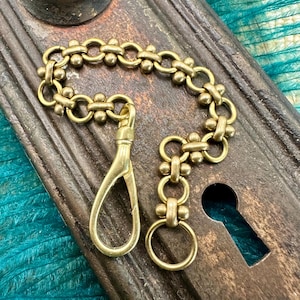 May include: A vintage brass chain with a clasp and ring, likely for a pocket watch or similar item. The chain is a warm gold color, with a decorative link pattern. The background is a weathered, dark metal surface.