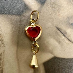 May include: A gold-colored charm with a heart-shaped red gemstone and a teardrop-shaped pendant. The charm is suspended from a small loop, and the heart has a gold border. The teardrop pendant is also gold.