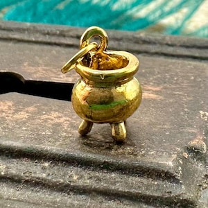 May include: A gold-colored metal charm in the shape of a cauldron with three legs. The charm has a loop at the top for attaching to a necklace or bracelet.