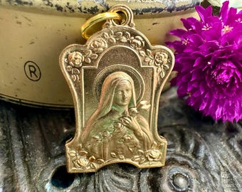 May include: A gold-colored religious pendant featuring a detailed relief of a woman with a halo, surrounded by roses. The pendant has a loop for hanging and is set against a blurred background with a purple flower.