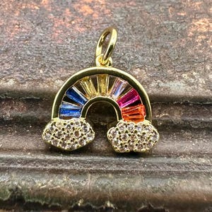 May include: A gold-colored rainbow pendant with a loop at the top. The rainbow features blue, yellow, orange, and pink rectangular gems. The clouds at the bottom are encrusted with small, clear gems.