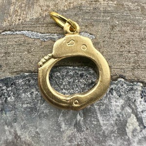 May include: A gold-colored handcuff charm with a small loop for attaching to a necklace or bracelet. The charm is detailed with rivets and a locking mechanism. The background is a textured, neutral surface.