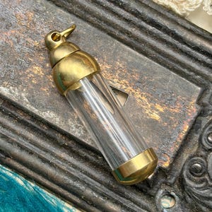 May include: A small, clear glass vial with brass-coloured metal accents. The vial has a rounded top and bottom, with a small loop at the top. The vial is empty and appears to be vintage or antique.