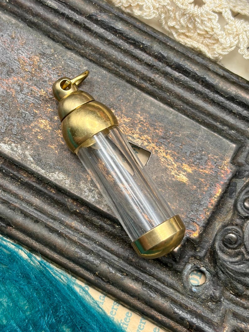 May include: A small, clear glass vial with brass-coloured metal accents. The vial has a rounded top and bottom, with a small loop at the top. The vial is empty and appears to be vintage or antique.
