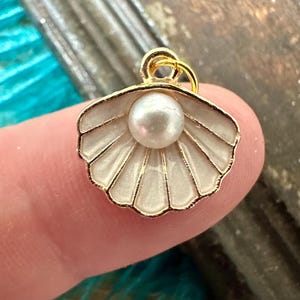May include: A gold-tone charm featuring a white seashell with a pearl inside. The charm has a loop at the top for attaching to a necklace or bracelet.