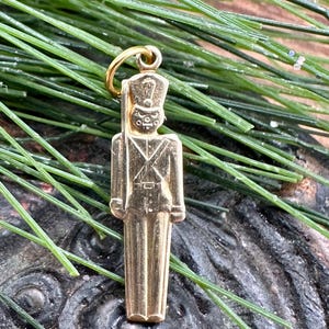 May include: A gold-colored charm in the shape of a toy soldier. The soldier is wearing a hat and has a rifle in its hand.