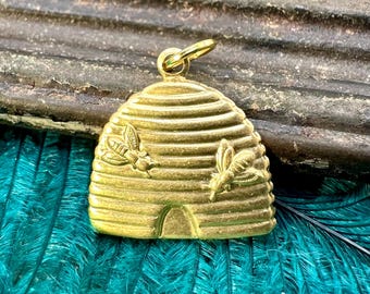 May include: A gold-coloured beehive charm with two bees on the front. The charm is detailed with a textured surface and a small opening at the bottom.