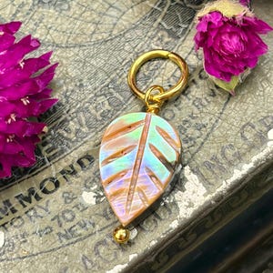 May include: A leaf-shaped pendant featuring an iridescent, multi-colored shell material with carved vein details. The pendant is attached to a circular gold-toned metal jump ring and includes a small gold-toned metal bead at the bottom tip.