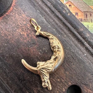 May include: A gold-toned pendant featuring a woman sitting on a crescent moon. The pendant is detailed with intricate carvings and has a loop at the top for attaching a chain.