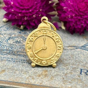 May include: A gold-coloured clock charm with a loop at the top. The clock face shows the time as approximately 1:30. The charm has a small base and is surrounded by purple flowers.