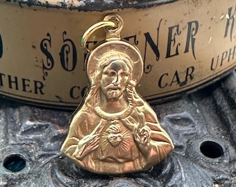 May include: A gold-colored pendant featuring a detailed depiction of Jesus Christ with a halo. The pendant shows Jesus with a heart and outstretched hands. The background includes a vintage tin with faded text.