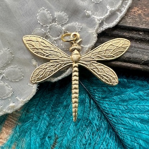 May include: A gold-colored dragonfly pendant with intricate wing detailing. The dragonfly has a textured body and wings, and a small loop for hanging. The pendant is set against a backdrop of white lace and teal feathers.