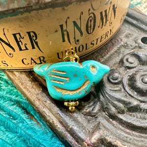 May include: A turquoise blue bird charm with gold accents. The bird is made of a textured material and has a detailed design. The charm is hanging from a gold chain.