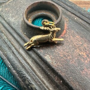 K&ouml;nnte beinhalten: Ein goldfarbener Hirsch-Anh&auml;nger mit detailliertem Design. Der Anh&auml;nger h&auml;ngt an einer Metallschaufel und liegt auf einer dunkelbraunen Oberfl&auml;che.