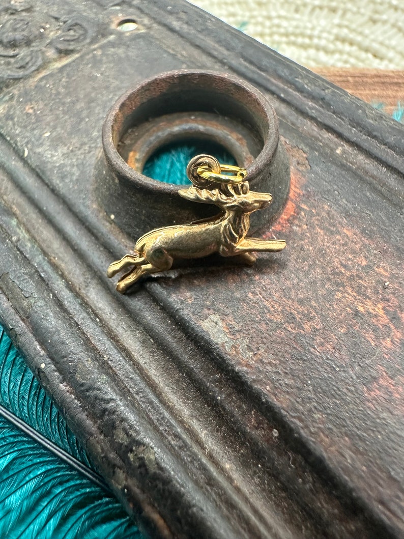 K&ouml;nnte beinhalten: Ein goldfarbener Hirsch-Anh&auml;nger mit detailliertem Design. Der Anh&auml;nger h&auml;ngt an einer Metallschaufel und liegt auf einer dunkelbraunen Oberfl&auml;che.