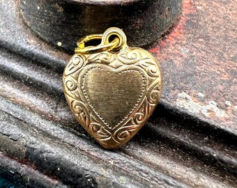 May include: A gold-toned heart-shaped charm with an ornate design. The charm has a loop at the top for attaching to a necklace or bracelet.