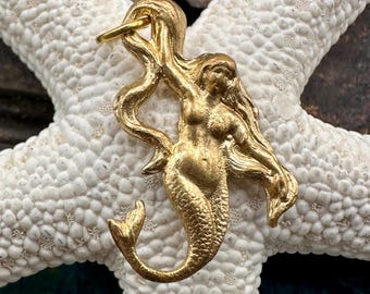 May include: A gold-coloured mermaid charm with a long flowing tail and hair. The charm is made of metal and has a loop at the top for attaching to a necklace or bracelet.