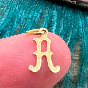 May include: Gold charm in the shape of the letter A. The letter has a decorative, ornate design.