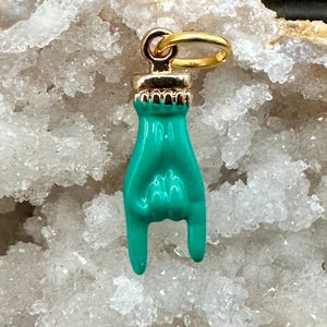 May include: A turquoise hand charm with a gold-colored bail, showing the 'rock on' hand gesture. The charm has a gold-colored crown-like detail at the top. The background is a white, crystalline surface.