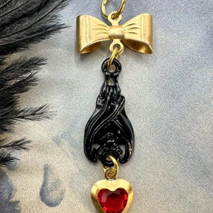 May include: A gold-colored bow pendant with a black bat charm and a red heart-shaped gem. The bat charm is suspended below the bow, and the heart is at the bottom. The pendant is designed for jewelry.
