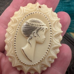 May include: A white cameo brooch with a detailed design of a woman's profile. The cameo is surrounded by a silver beaded border and a scalloped edge.