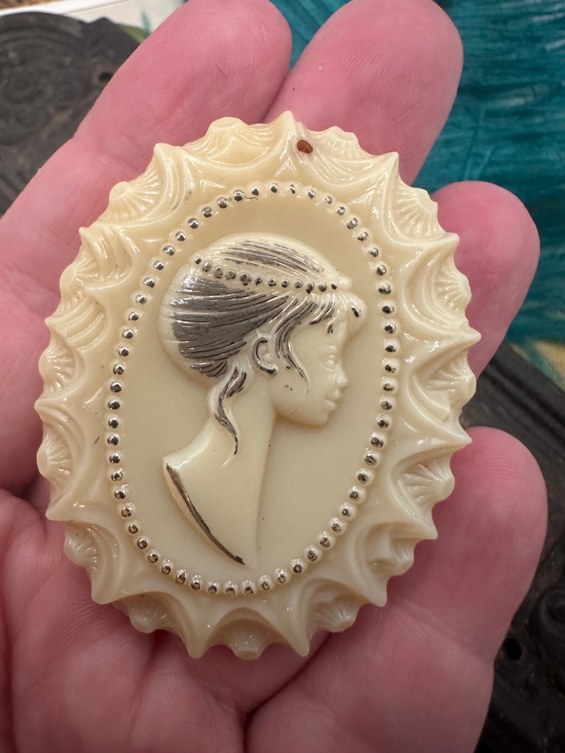 May include: A white cameo brooch with a detailed design of a woman's profile. The cameo is surrounded by a silver beaded border and a scalloped edge.
