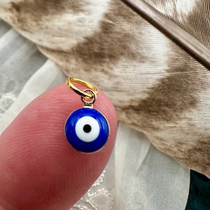 May include: A small gold charm with a blue evil eye design. The charm is on a finger.