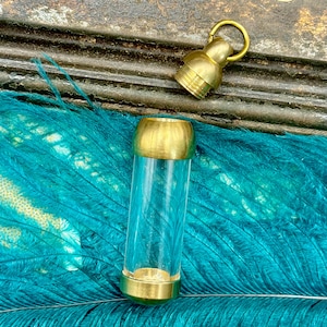 May include: A clear glass vial with gold-coloured metal ends and a matching gold-coloured cap. The vial is cylindrical and rests on teal feathers. A gold-coloured metal bell-shaped charm is also present.