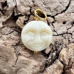 May include: A cream-colored pendant with a smiling face carved into it. The pendant has a gold-colored bail and a small gold-colored bead at the bottom. The pendant is round and has a smooth, glossy finish. The background is a textured, brown surface.