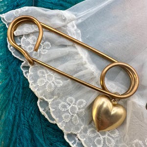 May include: A large gold safety pin with a heart-shaped charm attached. The pin is lying on a white lace fabric with a teal feather background.