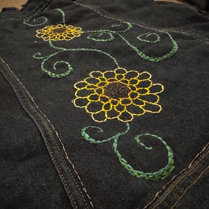 Può includere: Giacca di jeans blu scuro con ricami di girasoli gialli e tralci verdi. Il motivo floreale è cucito sul retro della giacca. La giacca presenta cuciture a vista e un classico look denim.