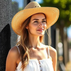 Puede incluir: Un sombrero de sol de paja con ala ancha. El sombrero es de color marr&oacute;n claro y tiene una textura tejida. La persona lleva una camiseta blanca con tirantes finos.