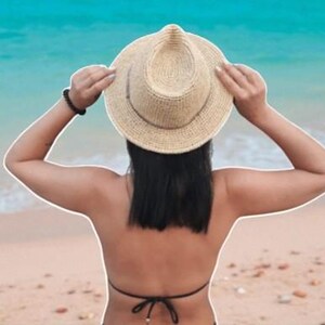 Puede incluir: Un sombrero de sol de paja con una corona redondeada y un ala ancha. El sombrero lo sostiene una persona con cabello oscuro, de pie en una playa de arena con el oc&eacute;ano de fondo. El sombrero es de color neutro.