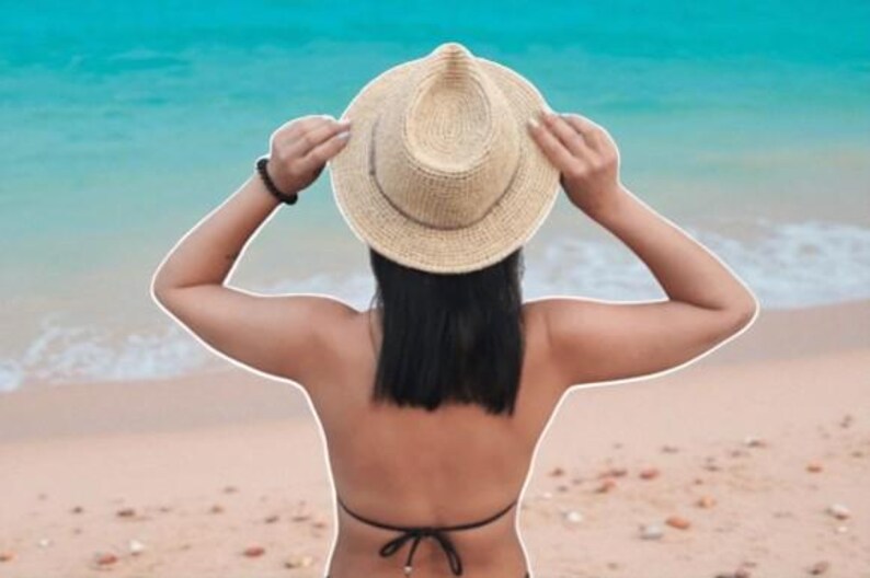 Puede incluir: Un sombrero de sol de paja con una corona redondeada y un ala ancha. El sombrero lo sostiene una persona con cabello oscuro, de pie en una playa de arena con el oc&eacute;ano de fondo. El sombrero es de color neutro.