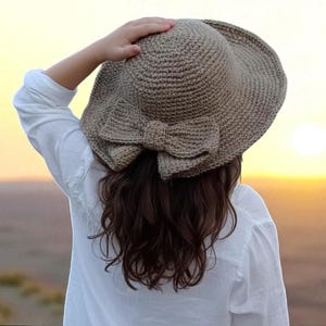 May include: A crocheted sun hat with a large bow. The hat is a light brown color and has a wide brim. The bow is centered on the back of the hat. The person is wearing a white shirt.