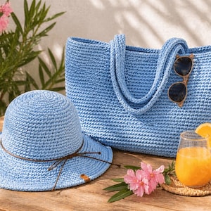May include: A light blue crochet tote bag, sun hat, and sunglasses rest on a wooden surface. A glass of orange juice with a slice of orange and a small coaster are also present. Pink flowers and green leaves are in the background.