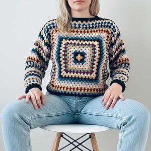May include: A multi-coloured crochet sweater with a square pattern, black cuffs and neckline. The central square design features teal, yellow, and burgundy, with a repeating pattern in various colours. The person is wearing light blue jeans.