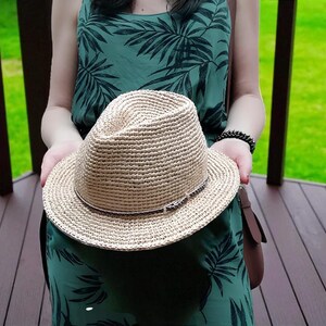 Puede incluir: Un sombrero fedora de paja tejida con una banda marr&oacute;n se sostiene frente a un vestido verde con un estampado de hojas tropicales. El sombrero tiene una corona cl&aacute;sica en forma de l&aacute;grima y un ala ancha. El sombrero es de color neutro.