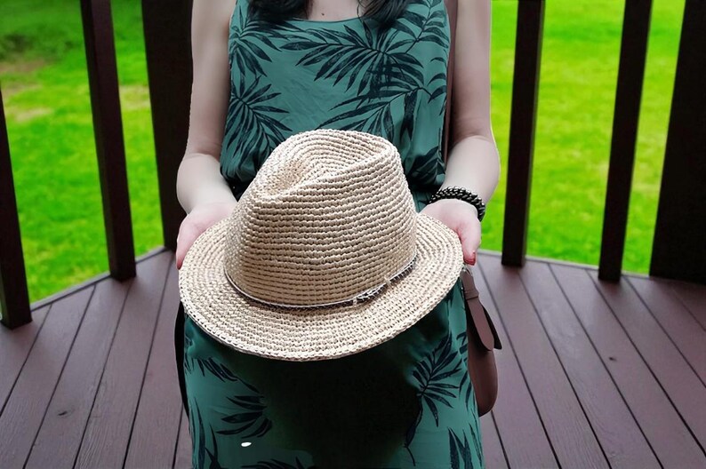 Puede incluir: Un sombrero fedora de paja tejida con una banda marr&oacute;n se sostiene frente a un vestido verde con un estampado de hojas tropicales. El sombrero tiene una corona cl&aacute;sica en forma de l&aacute;grima y un ala ancha. El sombrero es de color neutro.
