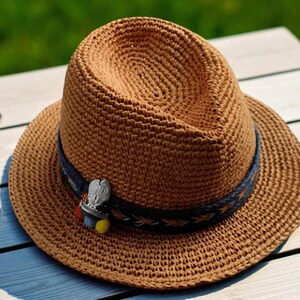 Puede incluir: Un sombrero fedora tejido marr&oacute;n con una banda decorativa y un peque&ntilde;o alfiler. El sombrero est&aacute; sobre una superficie de madera de color claro junto a unas gafas de sol con lentes oscuras. El sombrero tiene un dise&ntilde;o cl&aacute;sico y elegante.