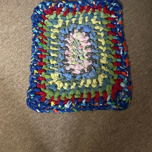 May include: A rectangular rag rug constructed from braided fabric strips in a multi-colored pattern. The center features light pink, gray, and blue tones, surrounded by concentric layers of green, red, and a thick outer border of dark blue fabric.