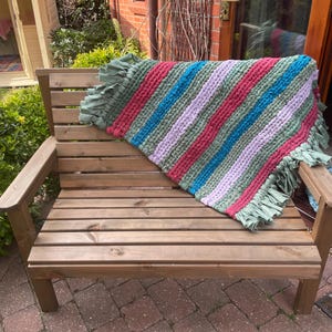 May include: A wooden garden bench with a handmade rag rug. The rug has stripes of green, red, blue, and lilac. The bench is made of brown wood and is in a garden setting.