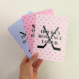 May include: Three rectangular cards featuring a repeating pattern of small hearts. Two cards are light pink with red hearts, and one is light blue with white hearts. Each card displays the black text HOCKEY ROMANCE LOVER over two crossed hockey sticks.