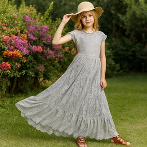 Puede incluir: Una persona joven con un vestido de encaje gris con dobladillo con volantes y mangas cortas, sandalias marrones y un sombrero de sol beige. El vestido tiene un patrón de encaje floral. El fondo presenta hierba verde y flores coloridas.