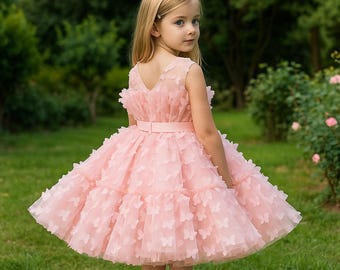 May include: A light pink dress with butterfly accents. The dress features a V-neck, a fitted waist with a matching belt, and a layered skirt. The dress is sleeveless and appears to be made of tulle. The background is a green garden.