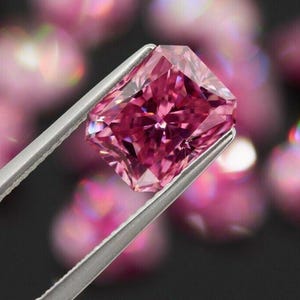 Può includere: Un diamante rosa acceso, taglio rettangolare, è tenuto da pinzette d'argento. Le sfaccettature della gemma riflettono la luce, mostrando il suo taglio brillante. Lo sfondo presenta sfere rosa sfocate, che esaltano il diamante. Questa immagine mette in risalto una pietra preziosa.
