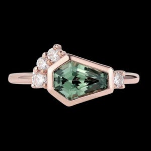 Könnte beinhalten: Ein roségoldener Ring mit einem geometrischen grünen Edelstein in der Mitte, flankiert von kleineren weißen Edelsteinen. Der Ring hat ein einzigartiges, modernes Design vor schwarzem Hintergrund.