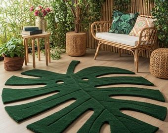 May include: A dark green, leaf-shaped rug dominates the foreground. The rug is a detailed replica of a Monstera leaf, with cut-out holes and textured detailing. The rug is placed on a light wood floor, with a tropical-themed outdoor setting in the background.