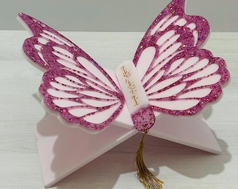 Könnte beinhalten: Ein rosa Schmetterling-Buchständer mit Glitzerakzenten. Die Flügel sind mit rosa Glitzer und Weiß umrandet. Ein kleines weißes Etikett mit arabischem Text ist in der Mitte befestigt, und eine goldene Quaste hängt darunter. Der Ständer ist hellrosa.