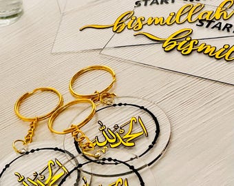 Könnte beinhalten: Klare Acryl-Schlüsselanhänger und -Anhänger mit goldfarbenen Buchstaben und Akzenten. Die Schlüsselanhänger haben goldfarbene Ringe und die Anhänger tragen den Schriftzug "Start with Bismillah" und arabische Kalligraphie. Die Artikel liegen auf einer weißen Oberfläche.
